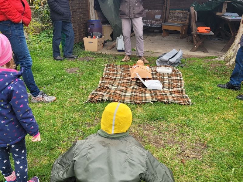 Man sieht mehrere Beine von Menschen, die um eine Wolldecke liegt, die auf der Erde liegt. Darauf ist ein Plastik-Oberkörper-Modell, welches zur Demonstration eines Defibrillators genutzt wird, zu sehen. Der Defibrillator ist angeschlossen.