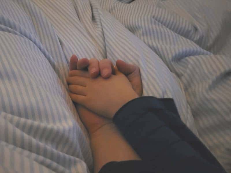 Eine große Hand und eine Kinderhand halten einander auf einer Bettdecke liegend.