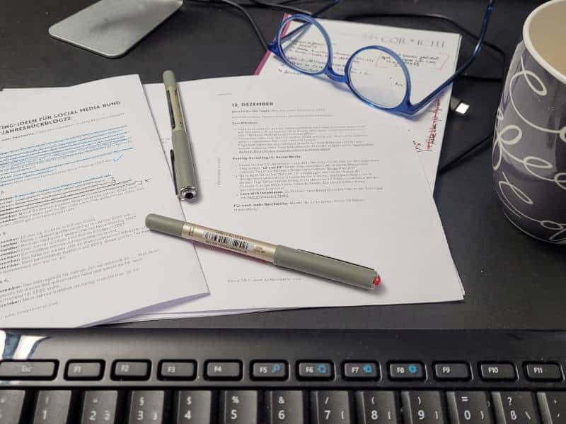 Ein paar Zettel, Stifte, eine Brille, eine Tasse und Teile einer Tastatur auf einem Schreibtisch.
