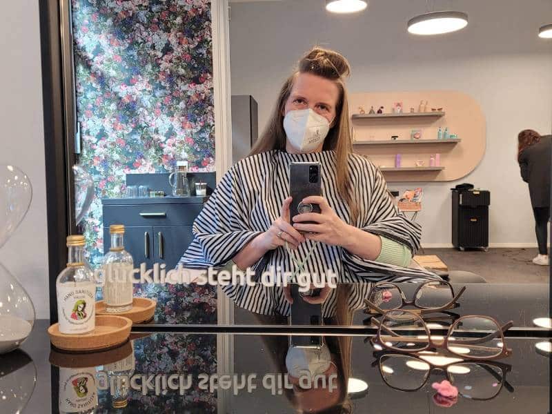 Spiegelselfie einer Frau im Friseursalon. Sie trägt einen Umgang und eine Maske. Ihre Haare sind oben auf dem Kopf zu Rolle gedreht. Auf dem Spiegel steht ein Spruch: "glücklich steht dir gut"