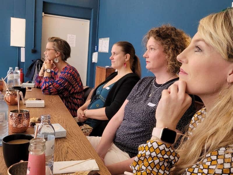 Vier Frauen sitzen an einem Tisch und schauen nach links. Sie hören jemandem zu, der nicht im Bild sichtbar ist.