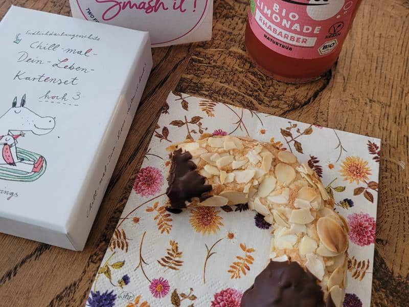 Auf einem Tisch liegt ein Mandelhörnchen auf einer Serviette. Daneben liegt ein Kartenset mit kleinen Zeichnungen und Sprüchen. Am oberen Bildrand ist noch eine Flasche Brause zu erkennen.