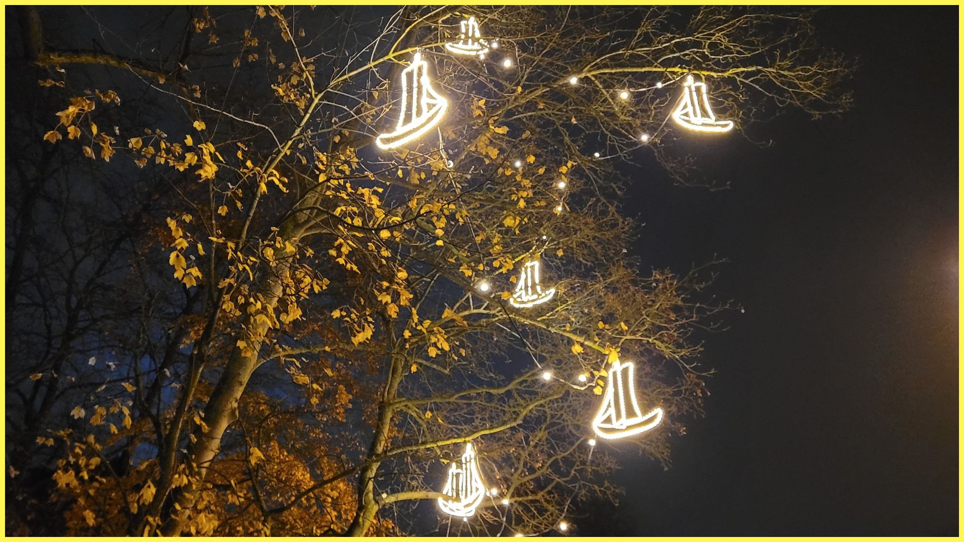 Blick auf einen Baum vor Nachthimmel, in dem beleuchtete Schiffe hängen. Diese Schiffe heißen Ewer.