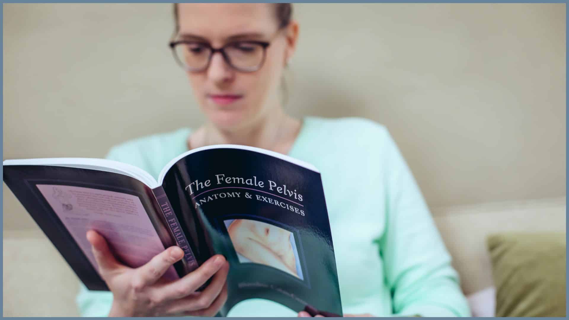 Eine Frau liest ein Buch mit dem Titel "The female pelvis - Anatomy and Exercises". Das Buch im Vordergrund ist scharf, die Frau im Hintergrund unscharf, aber erkennbar.