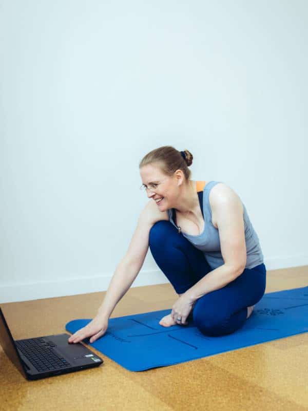 Eine Frau in Sportkleidung hockt auf einer Yogamatte vor einem Laptop.