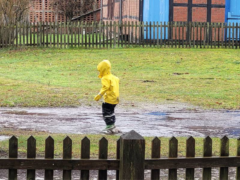 Man sieht auf einer Dorfstraße ein Kleinkind in Regenklamotten, was durch eine riesige Pfütze hüpft.