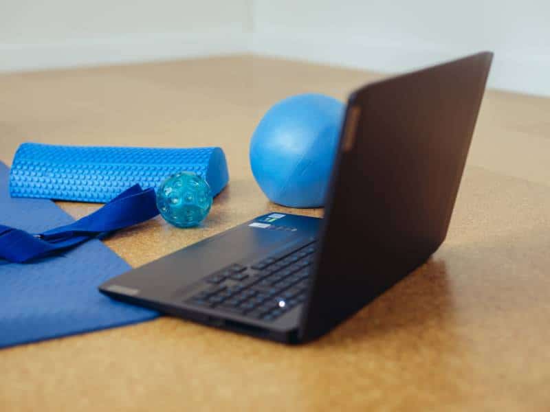 Ein aufgeklappter Laptop steht neben einer Yogamatte. Daneben liegen eine Halbrolle, ein Redondoball, ein Franklinball und ein Yogastrap. Alles Utensilien, die beim Online Beckenbodentraining zum Einsatz kommen.