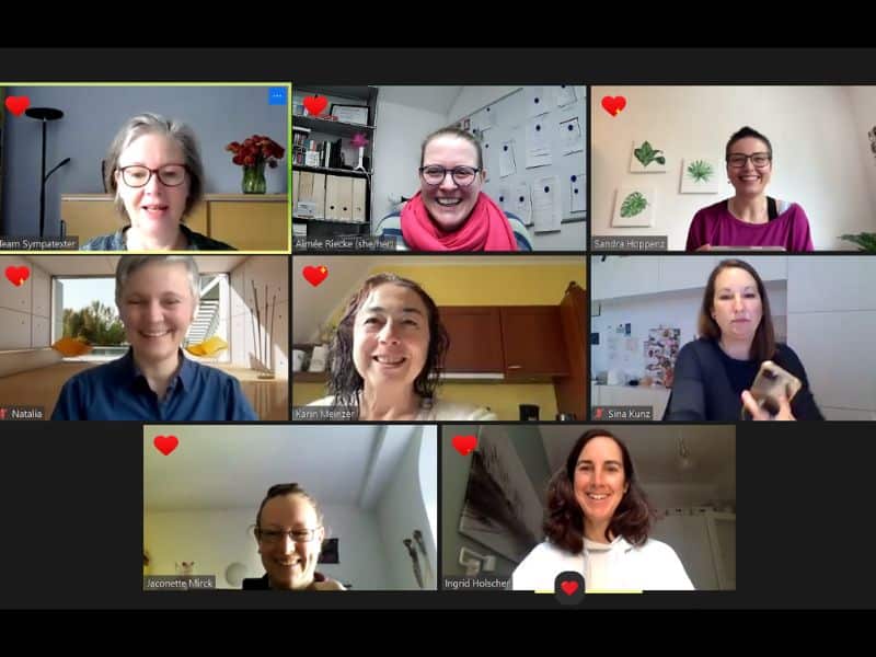 Ein Screenshot eines Zoomcalls mit 8 Frauen, von denen bei 7 ein rotes Herz im Bildschirm zu sehen ist. Alle lachen oder lächeln.