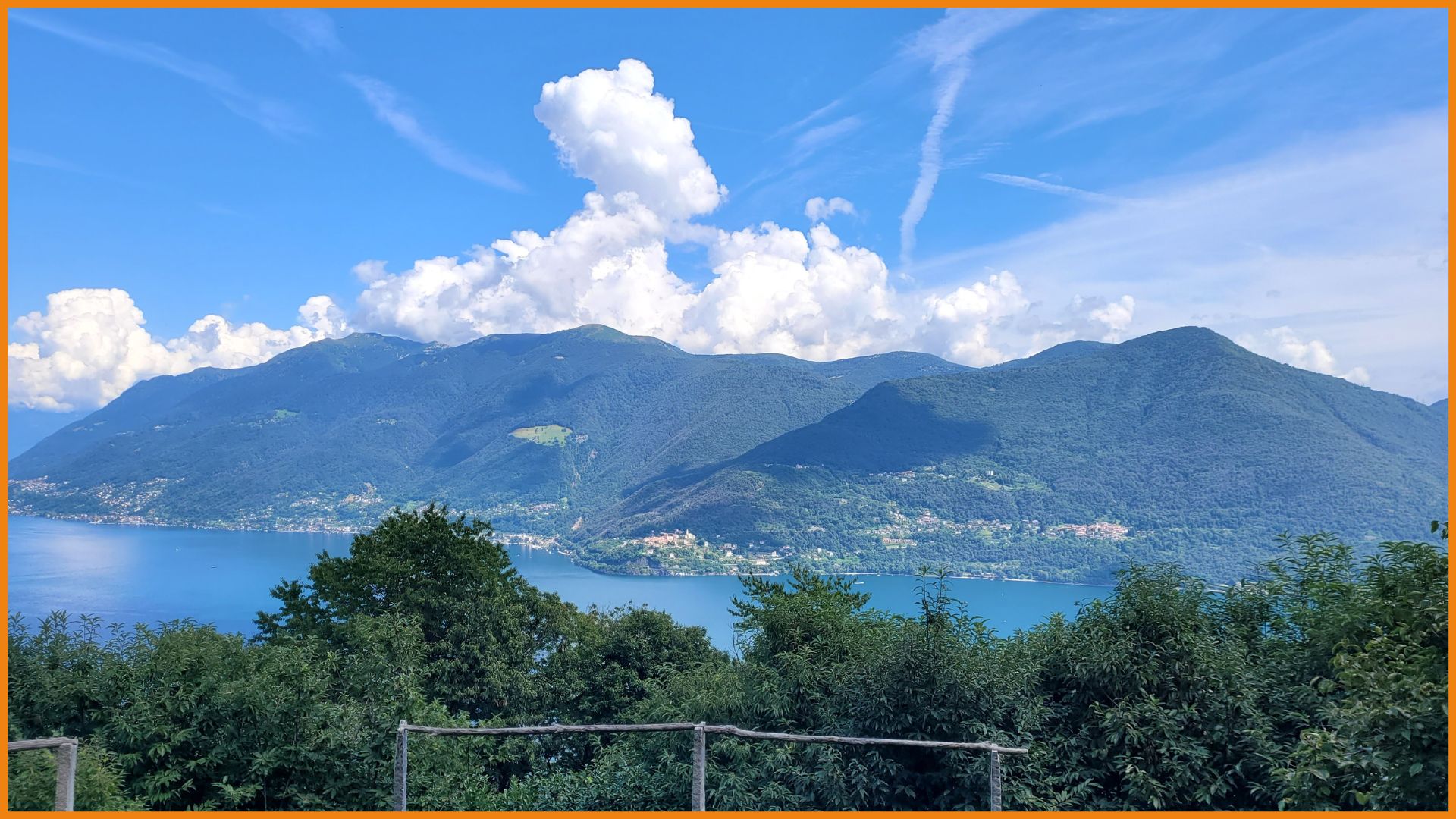 Blick über eine Reihe von Bäumen und einen See auf eine gegenüberliegende Bergkette. Der Himmel ist sehr blau, es ist sonnig und dein paar sehr weiße Wolken hängen hinter den Bergen.