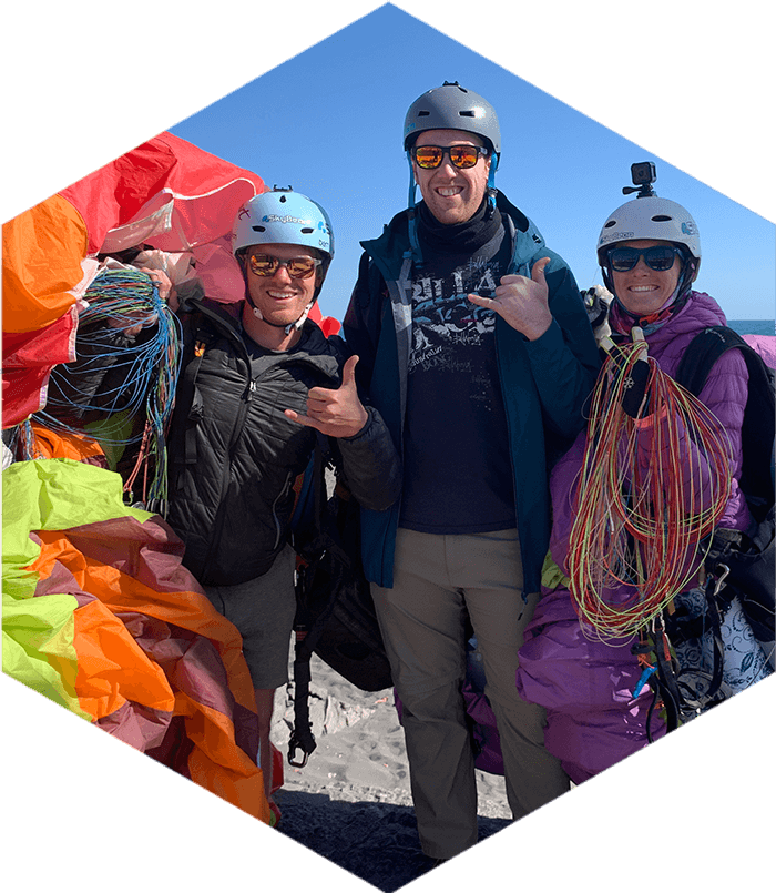 three smiling people wearing helmets and carrying paragliding kit