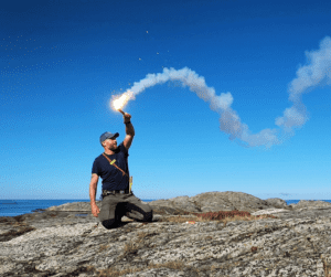 Torbjörn Selin sitter på en klippa med blå himmel bakom med ett tänt nödsignalbloss i handen. det lyser starkt och ryker.