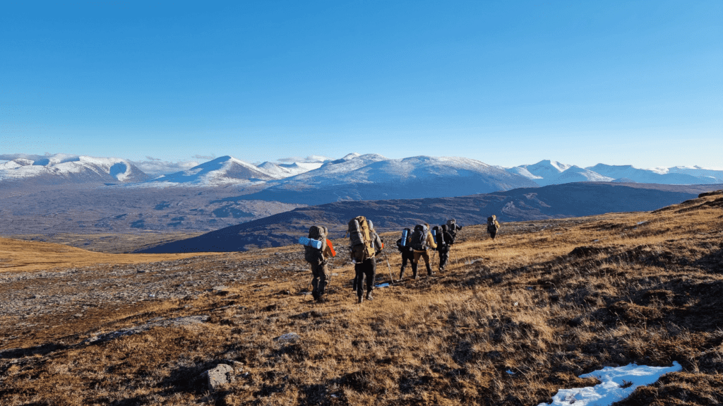 En grupp med ryggsäckar guidas bort från kameran över gräsbeklädd mark med snötäckta berg i bakgrunden.