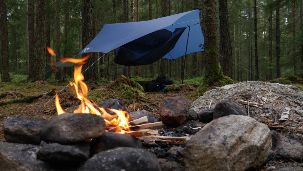 En fin lägereld i förgrunden. Längre bak hänger en hängmatta med tarp över. En del av allemansrättens rättigheter.