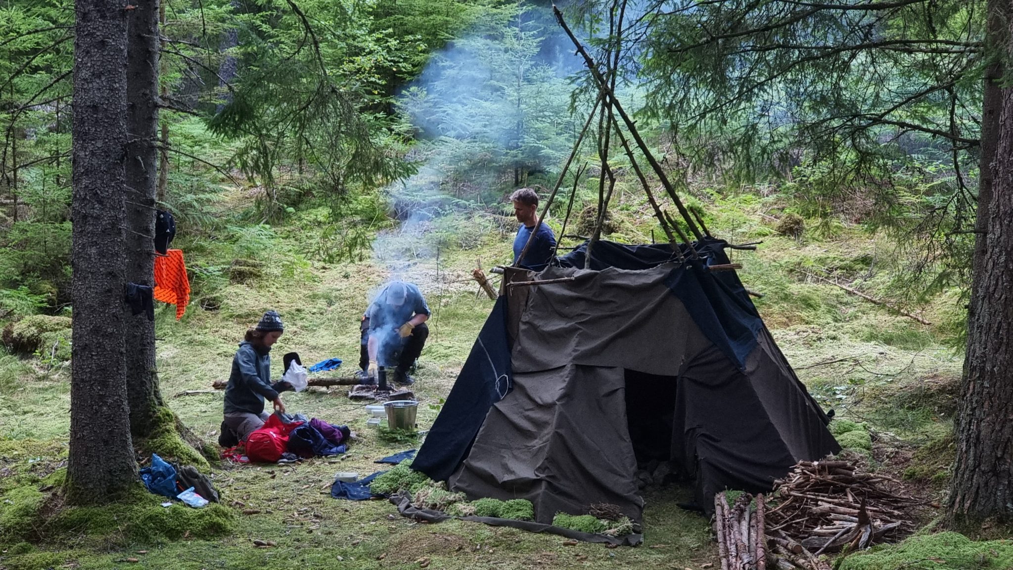 Eldhydda som liknar indianernas tipi i förgrunden, vid sidan tre personer från överlevnadskursen som packar en väska, eldar och en som bär ved. Det ryker från elden.