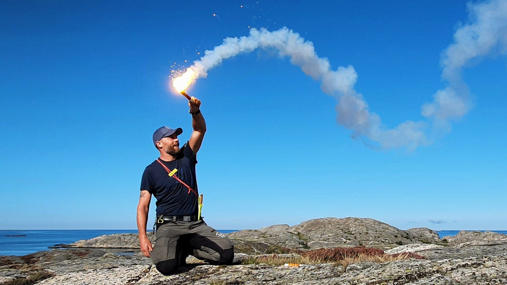 Torbjörn Selin sitter på en klippa med blå himmel bakom med ett tänt nödsignalbloss i handen. Det lyser starkt och ryker.