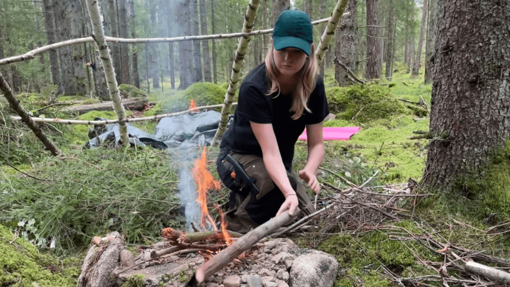 Ung tjej med grön keps lägger ved på elden. I bakgrunden byggs ett vindskydd.