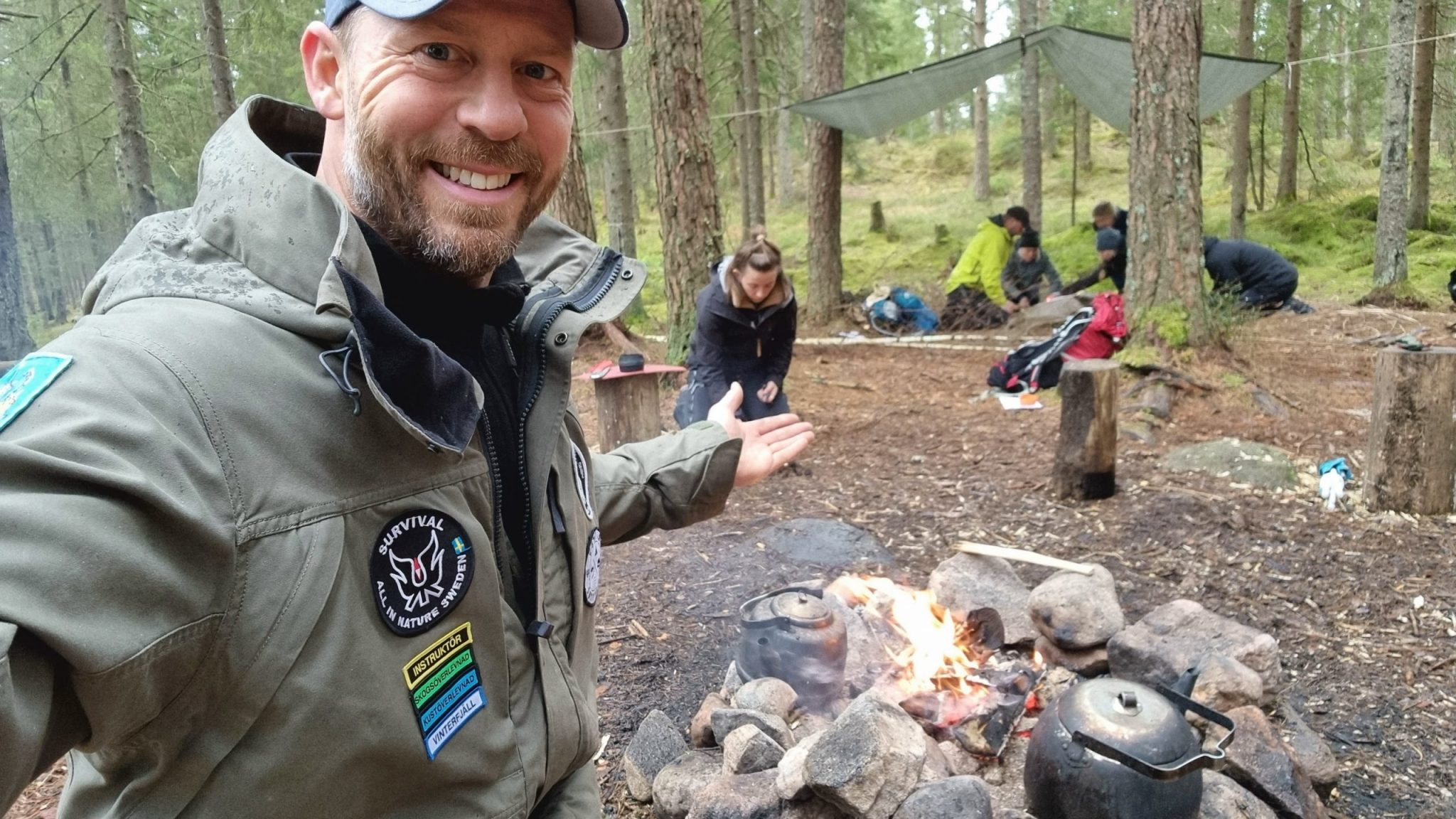 Torbjörn visar en välkomnande gest mot skogen en eld och en grupp människor som jobbar på.