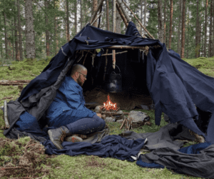 En man tittar på en eld i en bivack bygd av mörkblåa tyger, omgiven av skog.