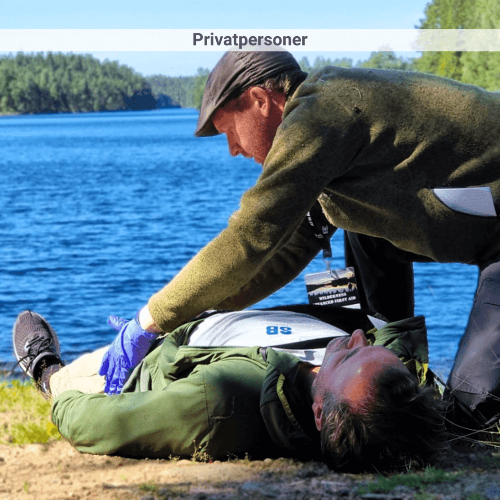 En man i grön tröja och blå plasthandskar undersöker en annan man som ligger på marken vid sjökanten.