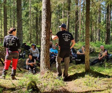 Instruktörer Torbjörn och Sanela står med ryggen mot kameran, friluftskläder, röda och beige byxor. Bortanför dem vända mot kameran sitter hela kursen och tittar upp mot dem. Vänster i bild ser man en eldhydda.