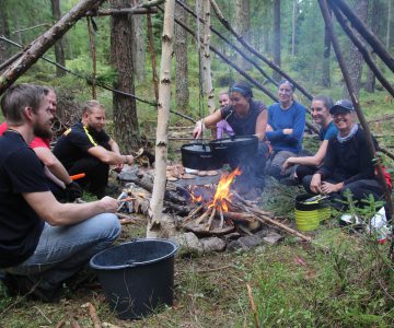 En teambuildinggrupp killar och tjejer lagar mat kring elden i sitt skydd i skogen ett event utöver det vanliga