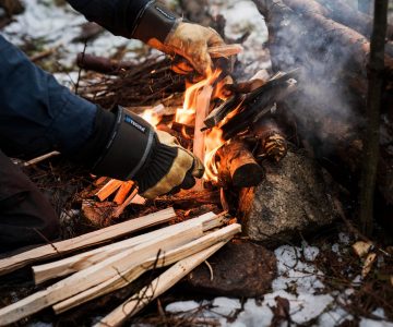 Torbjörn vårdar en lite större eld och lägger varsamt i pinnar snö på marken och handskar på händerna