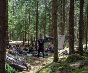 Instruktör Torbjörn Selin står vid en liten whiteboard i skogen och håller lektion under överlevnadskurs.