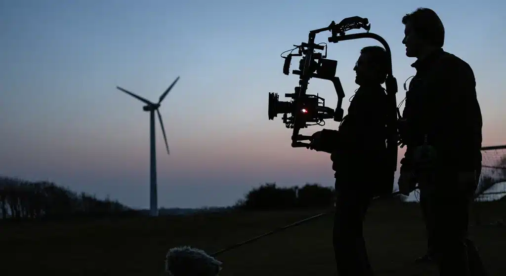 Silhouette of a film crew shooting at a field with a wind turbine in the background