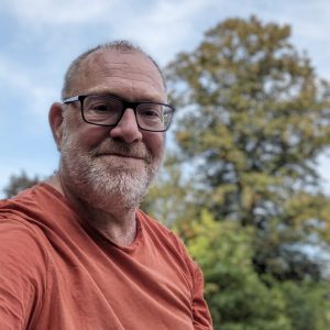 short haired middle aged person with glasses wearing a orange tee shirt