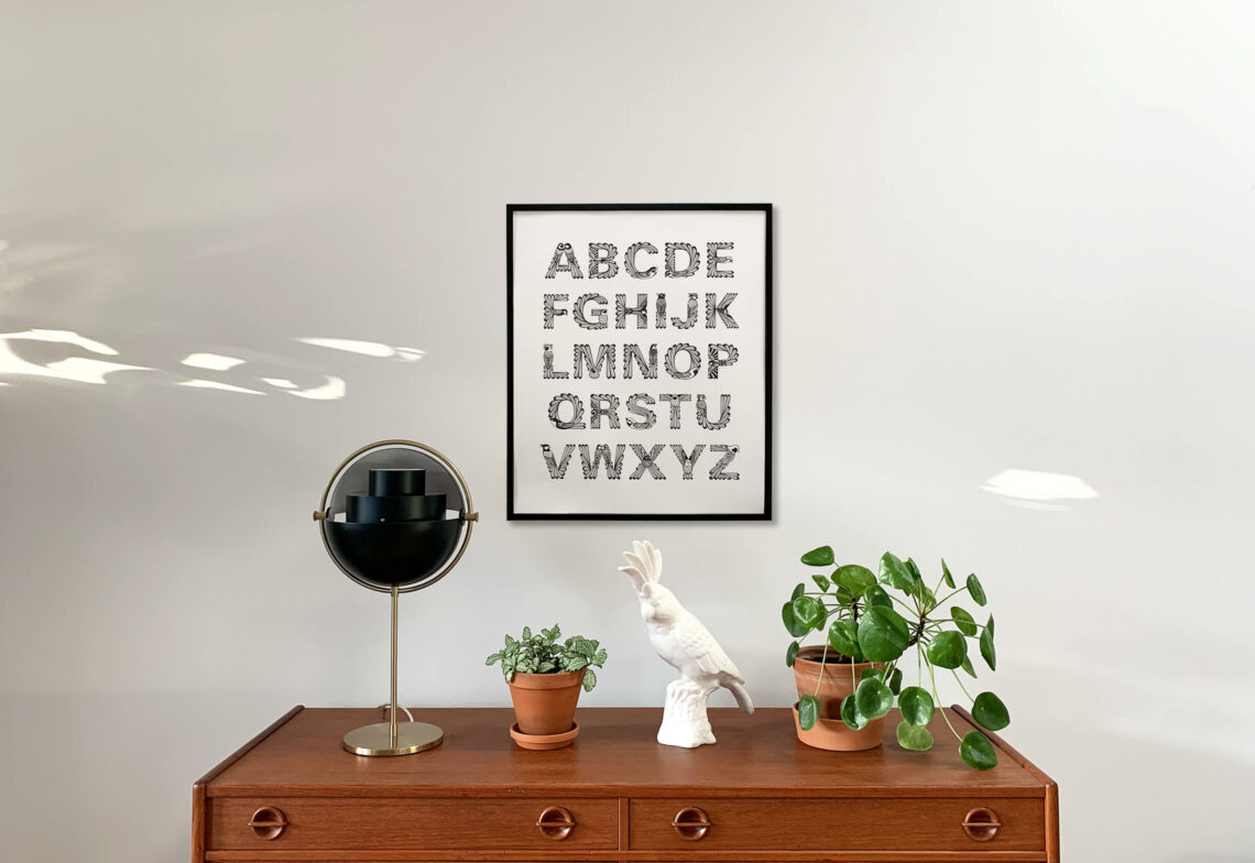 Inramad affisch av alfabetet "Ornis" hängandes på en vit vägg med en byrå, lampa, växter och prydnadsfågel under affischen