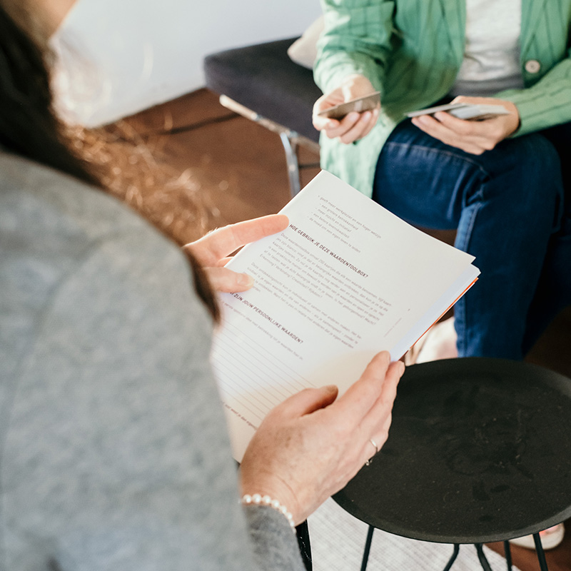 Een jobcoach is in gesprek met een patiënt. De coach heeft een boekje vast, de patiënt kaartjes van een opdracht.