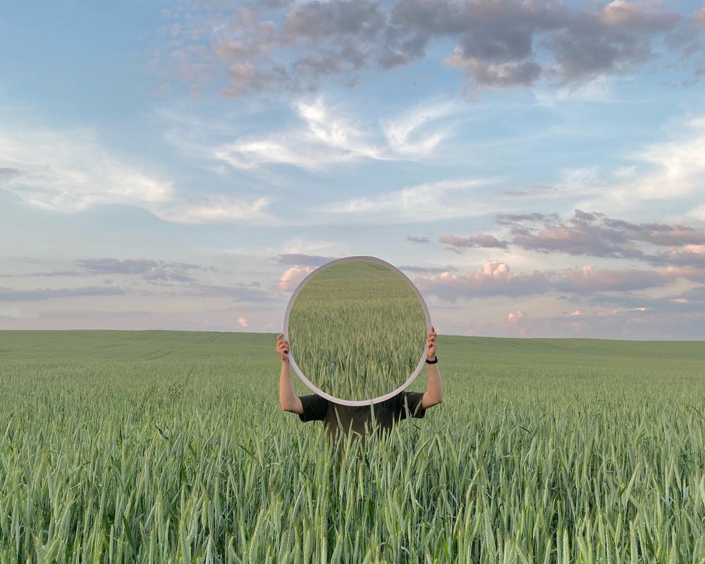 Iemand staat in een veld en houdt een spiegel voor zijn gezicht waardoor je enkel het graan in de spiegel ziet