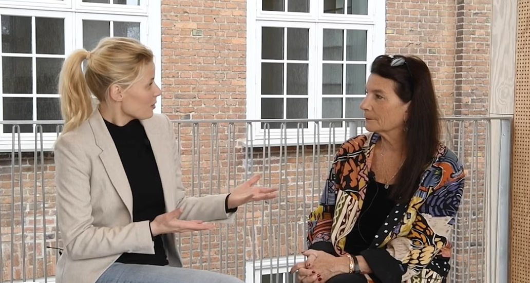 Module 3 Two women sit indoors in front of large windows and a brick wall and talk to each other. The woman on the left gestures with both hands while the other listens.