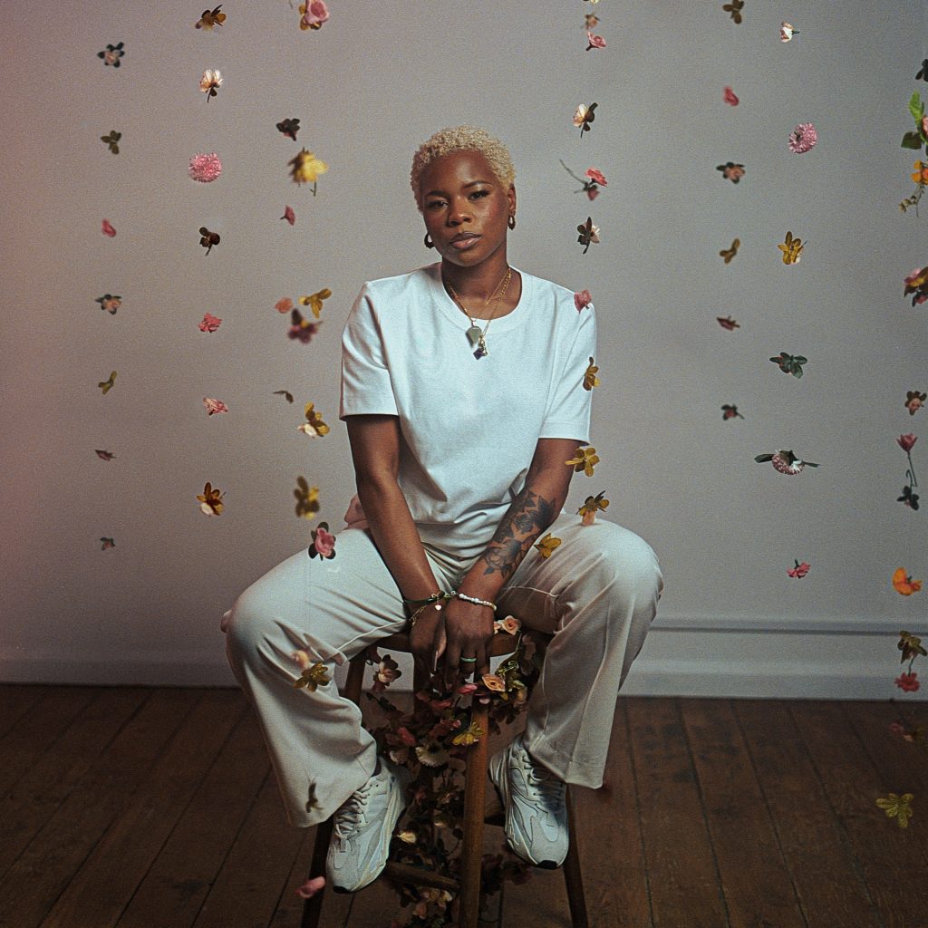 Laolu sat on a wooden stool with flowers wrapped around it on a wooden floor. The background is a plain white wall.