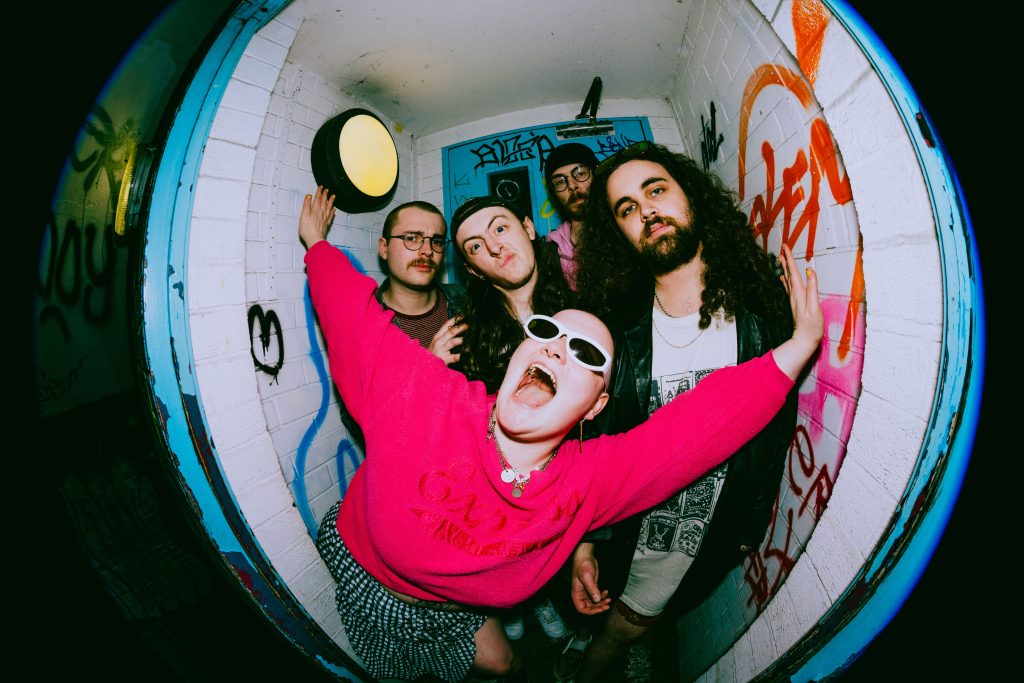 Gen and the Degenerates pictured in a stylised fishbowl lens standing in front of a blue door with graffiti all over it and the surrounding brick walls.