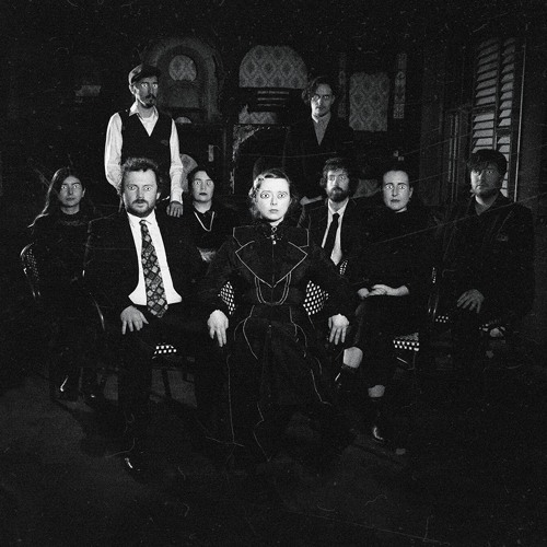 Black and white picture of people with straight faces sitting on chairs and two standing at the back with what appears to be drawn on eyes.