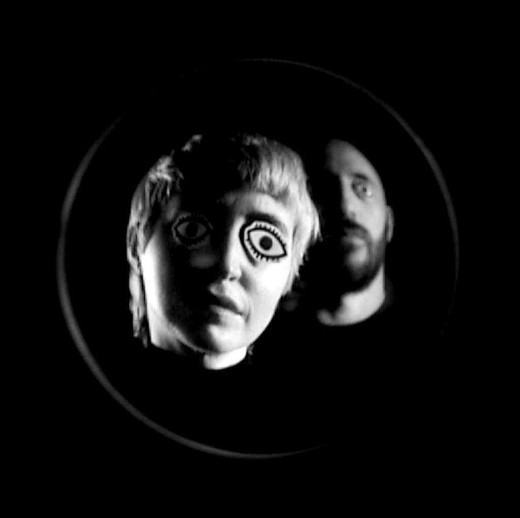Woman (foreground) and man (background) in a round frame on a black background. Paper cutouts with eyes darwn on, cover their eyes.