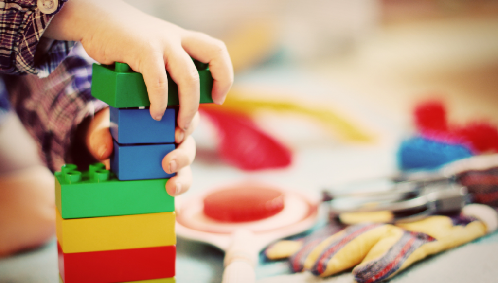 A close-up of a child's hand stacking colorful plastic building blocks on a play surface. The background is blurred and features various toys, including a paddle, gloves, and other play items. The scene suggests a creative and playful environment.
