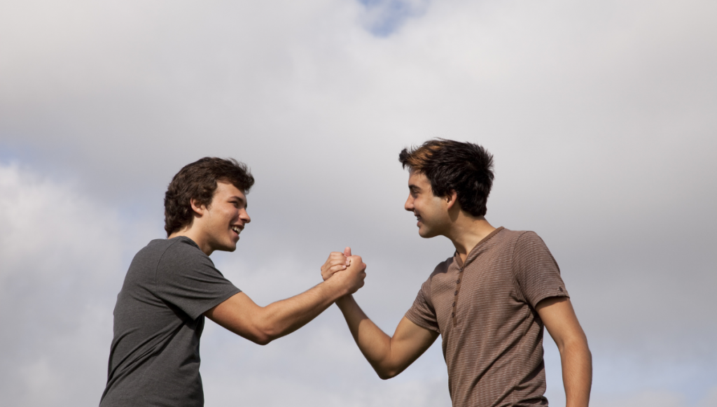 Two young men with light skin are smiling and shaking hands in an outdoor setting with a cloudy sky in the background. They appear to be engaged in a friendly and positive interaction.