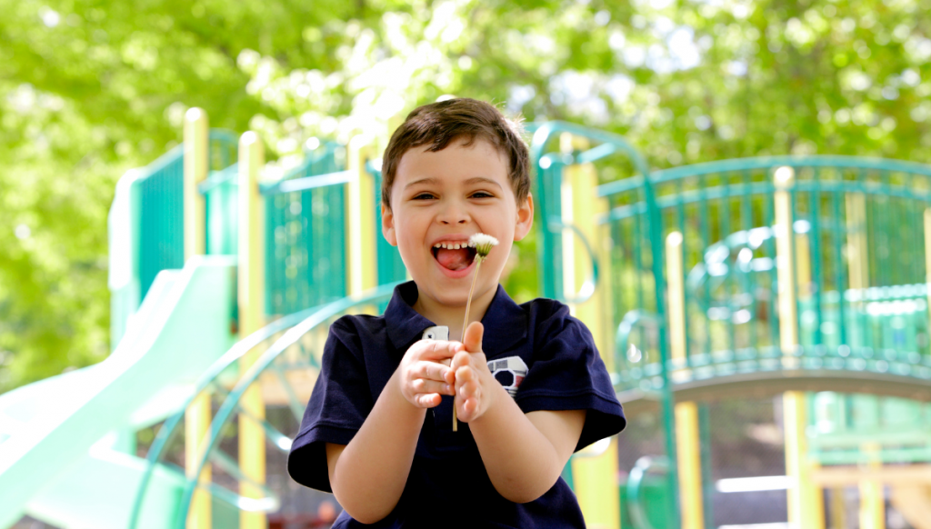 A young boy with short brown hair and light skin is smiling joyfully while holding a small white flower. He is wearing a navy blue polo shirt with a soccer ball emblem on the chest. Behind him, there is a colorful playground with green and yellow structures, including slides and climbing bars, surrounded by lush green trees. Sunlight filters through the leaves, creating a bright and cheerful atmosphere.