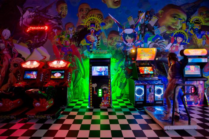 Several glowing arcade gaming machines stand in a room against a wall decorated with vivid gaming-related images