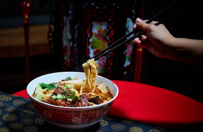 A hand holding chopsticks wrapped at the end in thick noodles lifted from a bowl of beef broth at Superfresh