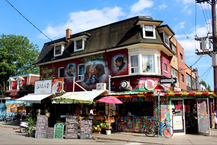 A red-brick corner building with a mural of three young people on high the wall. Beneath it is a stall called The Salad Shed