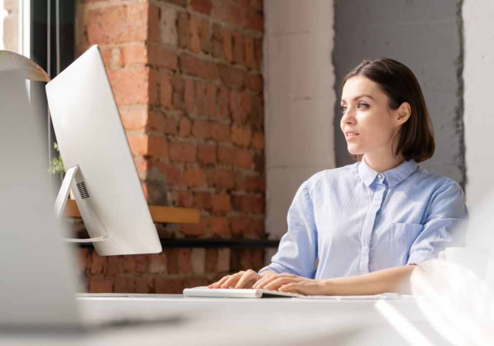 A young woman is pressing the keys of a computer keyboard while looking at the screen and working on the computer.