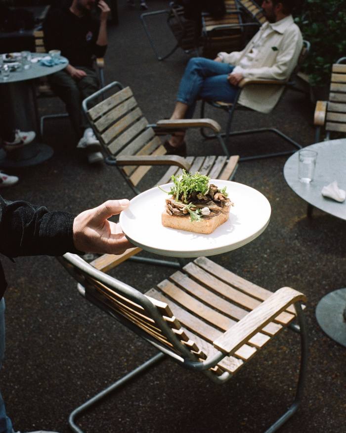 A man carrying a plate of roast mushrooms on a toasted homemade brioche bun, with black garlic aioli, Jerusalem artichoke ‘cream cheese’ and mizuna, on the terrace of Miró Manufactura de Café 