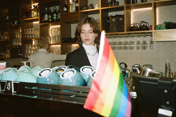 Two employees behind the counter at ViCafe’s branch inside LGBTQ+ space Kweer, with a rainbow flag in the foreground beside rows of blue coffee cups