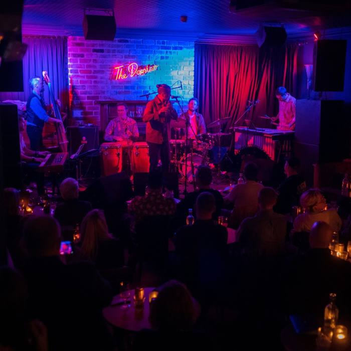 The interior of a bar or club, with a band on stage bathed in red and purple light, and the audience in darkness