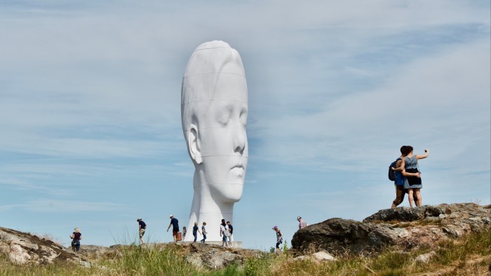 A gigantic white sculpture of a woman’s head with her eyes closed. In the foreground, human figures are dwarfed by the scale of the sculpture