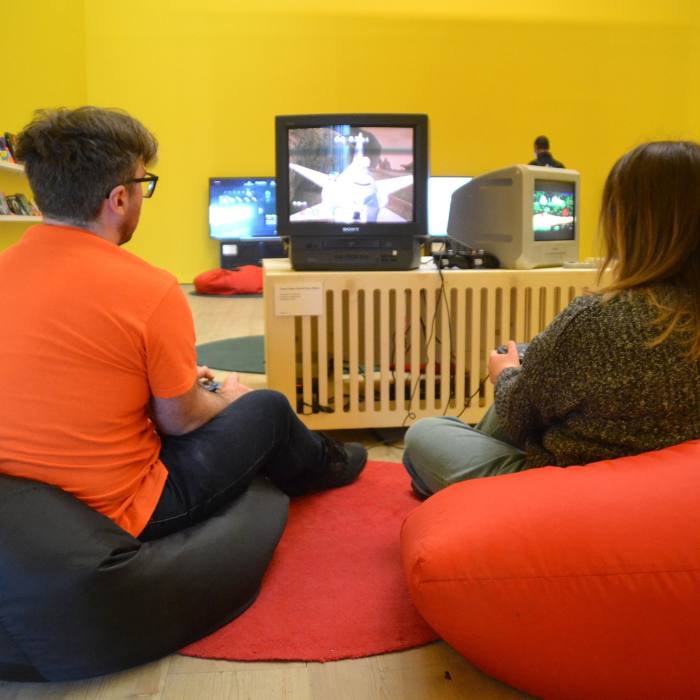 Two people sit on beanbags playing video games in a colourful room with several screens on low tables