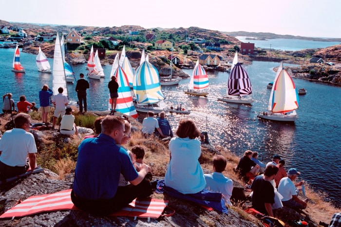 Spectators sit in the foreground watching as multiple sailing boats cluster in a harbour